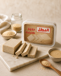 A clear plastic tub of El-Batal tahini paste with Arabic text on the label, displayed on a marble board with sliced beige paste, small bowls of sesame seeds, and a wooden spoon on a neutral beige background.