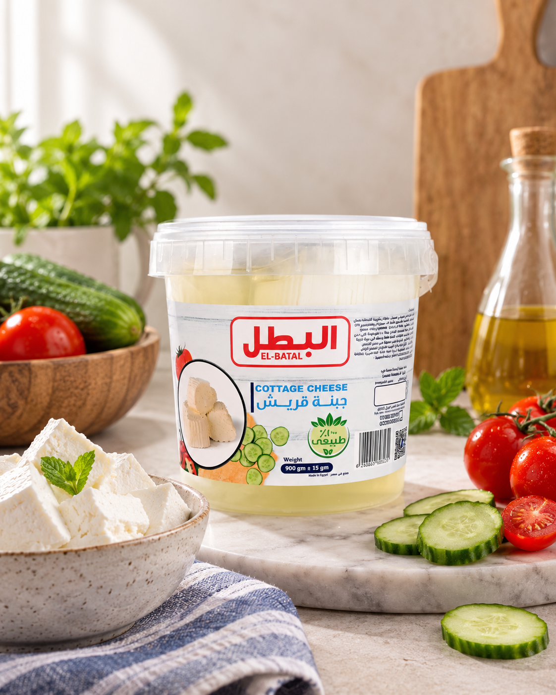 A tub of El-Batal Cottage Cheese placed on a marble surface with tomatoes, cucumber, herbs, and a glass bottle of oil around it, creating a styled kitchen scene.